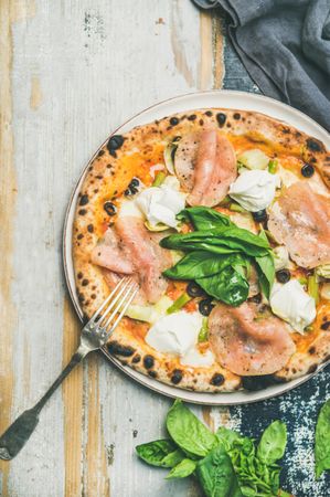 Freshly made wood fire oven pizza with spinach, ham and olives, on wood background with fresh herbs