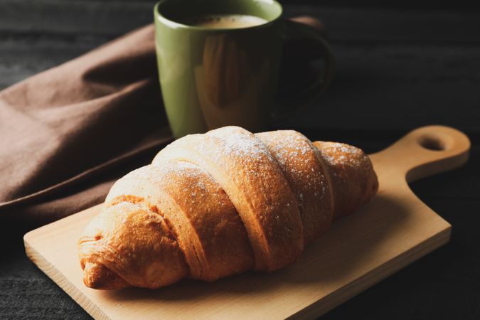 Board with croissant and mug of coffee on dark wooden background, close up