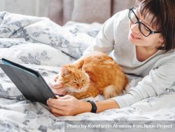 Cute ginger cat and woman in glasses lying in bed looking at tablet 49lJn5