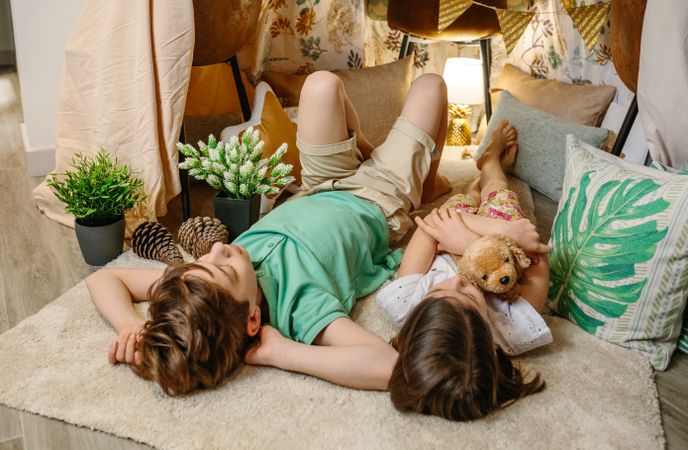 Serene boy and little girl embracing stuffed dog resting surrounded by cushions and plants in teepee