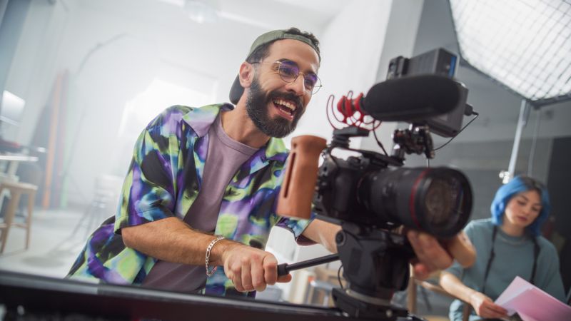 Young Male Cinematographer With Beard, Wearing Colorful Shirt, Joyfully Operates Professional Camera In Studio Setting, With Focused Female Colleague In Background Reviewing Notes.