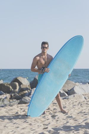 Male surfer holding surfboard standing in front of rocks near the coast