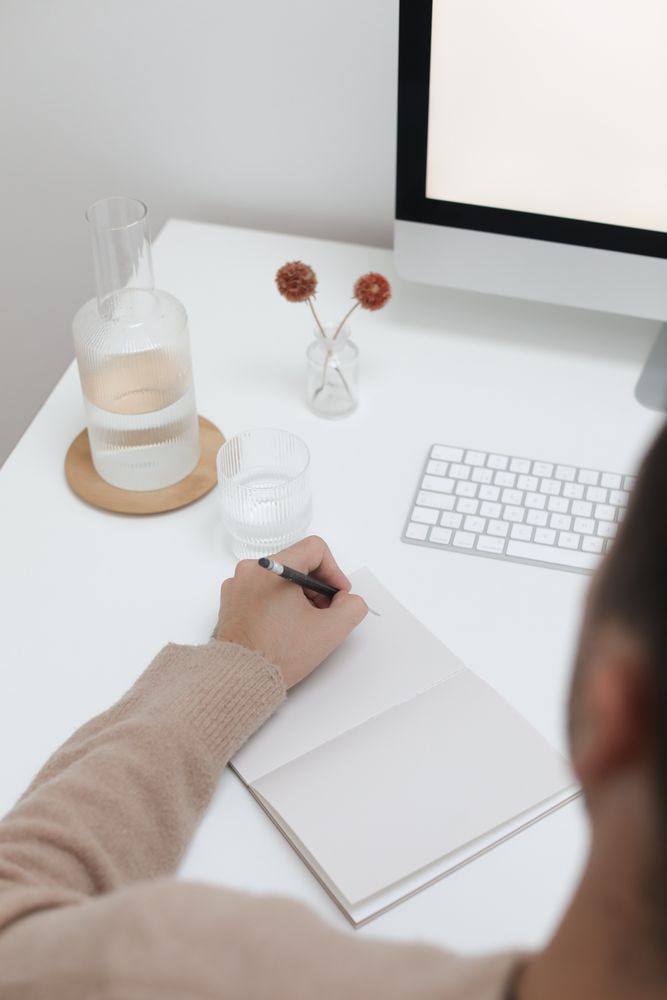 Person writing on notebook near computer desktop on light desk