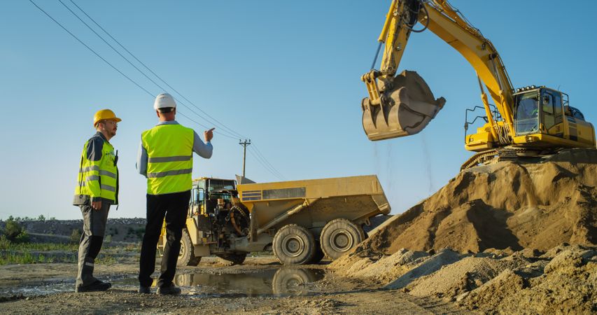Male Real Estate Investor And Project Manager Talking On Construction Site Of Apartment Block. Colleagues Discussing Building Progress, Business. Excavator Loading Sand In Industrial Truck.