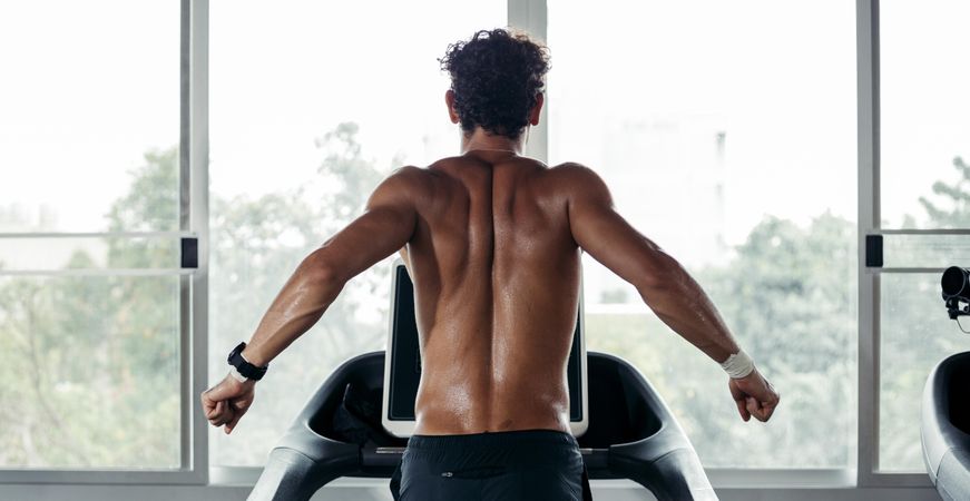 Muscular male’s back on treadmill near window