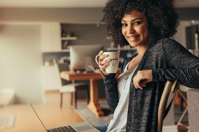 Portrait of attractive woman with curly hair having coffee at home with a laptop.