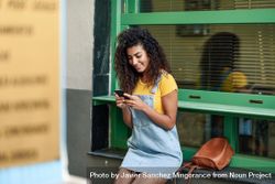Smiling Arab woman perched on green window sill wearing denim dress while texting, copy space 0LROA4