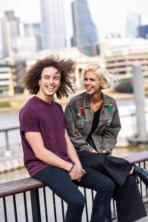 Smiling blonde female and curly haired male chatting while sitting on fence over river