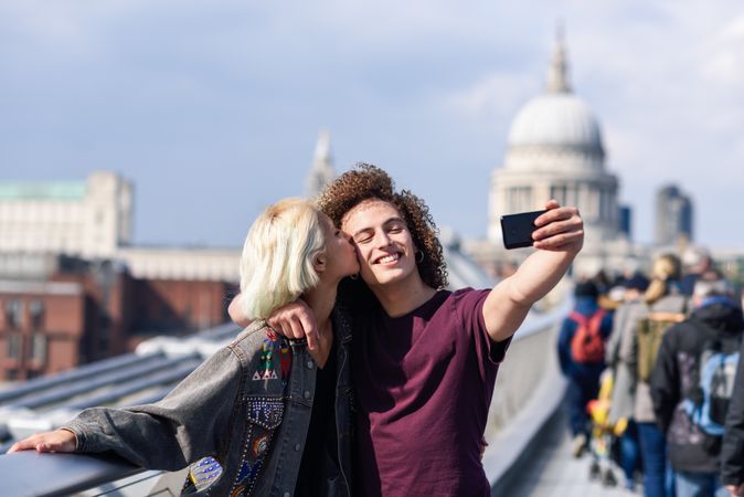 Blonde female and curly haired boyfriend taking picture with cell phone on bridge in London