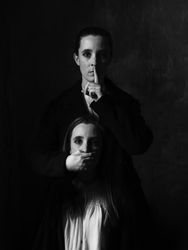 A young woman asks for silence with a finger gesture on her lips, while she covers the mouth of another woman, identical to herself, in a dark room