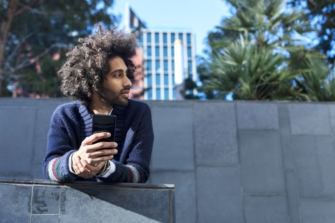 Black man in cardigan leaning forward outdoors on sunny day holding smartphone