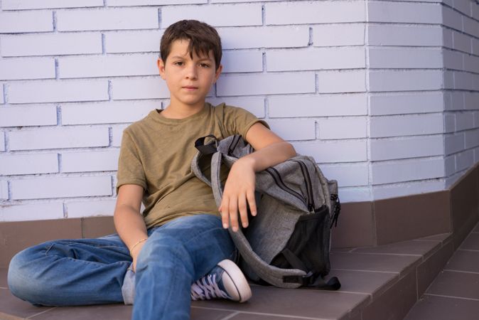 Boy sitting on ground in school corridor next to backpack