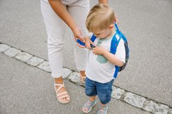 Unrecognizable mother helping child with backpack while standing on asphalt road in daylight