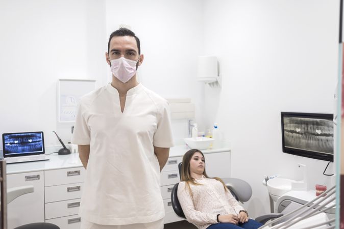 Male dentist in mask pictured in his office with teenage patient in background