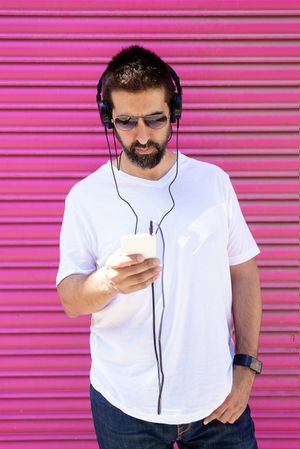 Latino man in headphones looking at phone in front of pink wall