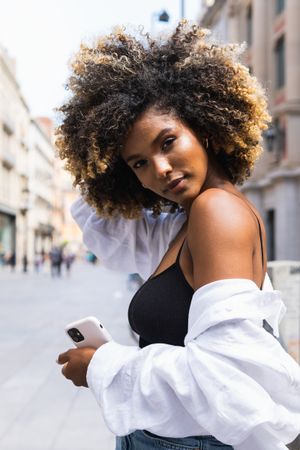 Black woman touching hair while standing on street with smartphone