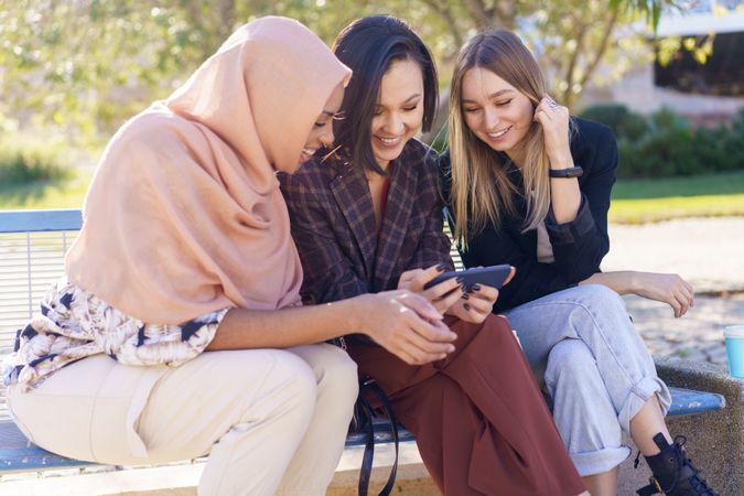 Happy women sitting on outdoor park bench discussing something on phone screen