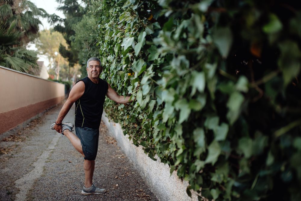 Mature man stretching leg holding botanical wall in street
