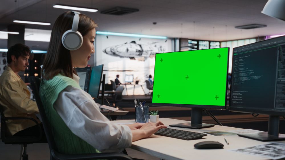 Female Asian Game Programmer Coding On Desktop Computer With Green Screen Chromakey On Display In Game Development Studio Office.