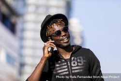 Smiling Black man wearing hat & sunglasses standing on the street talking on cellphone 41l28L