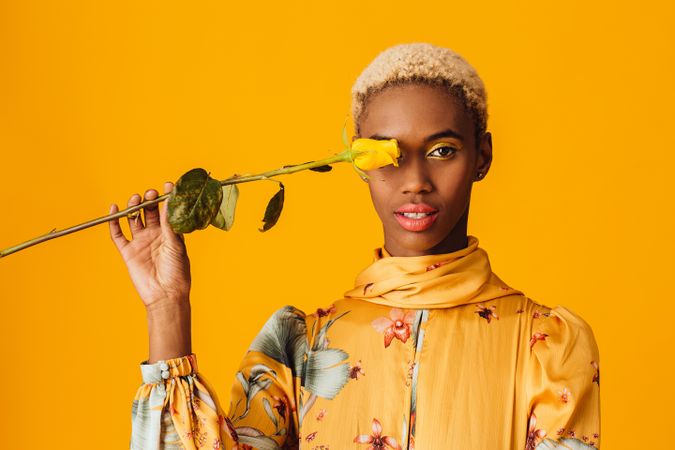 Black woman with short blonde hair holding yellow flower over eye