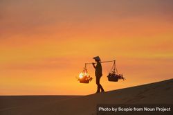 Silhouette of farmer with conical hat holding carrying pole walking on sand dune at sunset 47EkA0