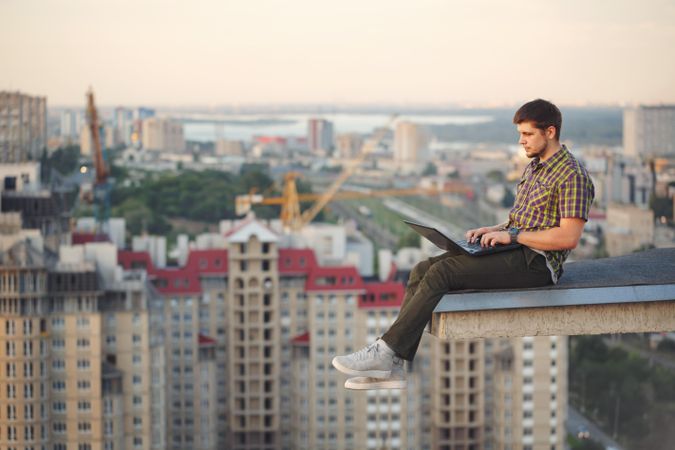 Man perched on roof with legs dangling over city while working on laptop