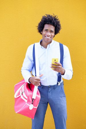 Happy male wearing suspenders and sports bag with phone in front of yellow wall