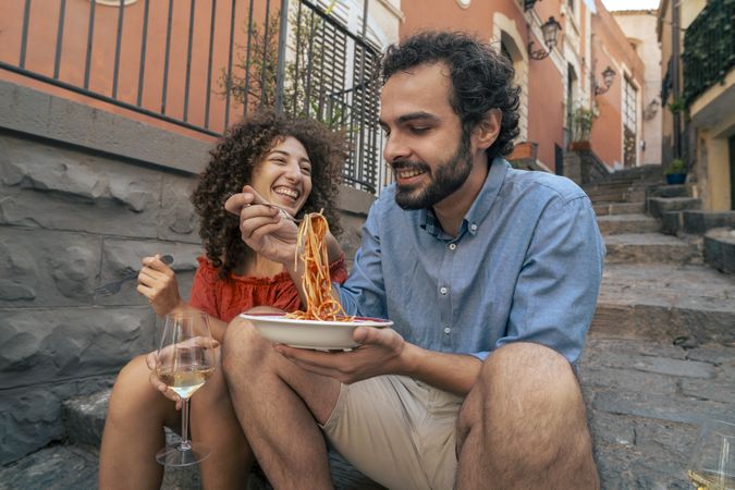 Playful young couple sharing one plate of pasta on city steps.