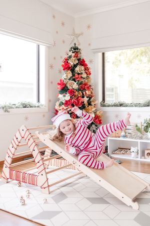 Girl on wooden playground slide beside Christmas tree in living room