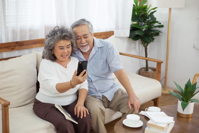 Happy Asian couple sitting on sofa using smartphone video chat