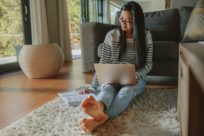Woman sitting on floor leaning to sofa looking at book with laptop on her lap