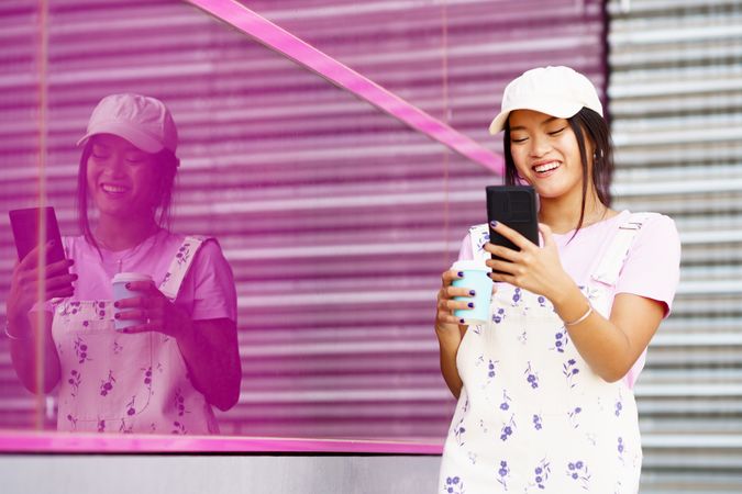 Laughing trendy Chinese woman walking outside in coveralls holding to go coffee and smart phone