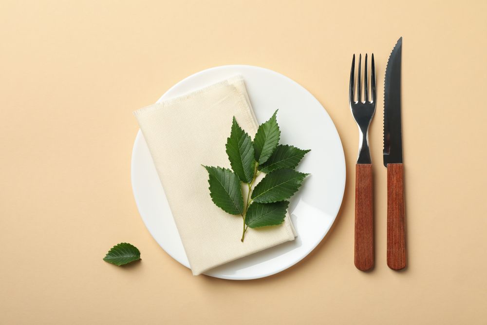 Plate with napkin, cutlery and leaf on beige background, top view
