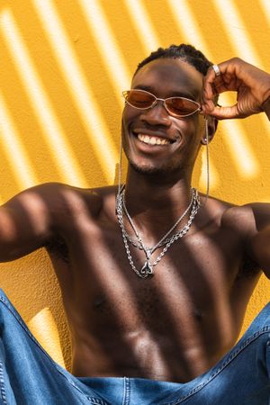 Cheerful Black man in trendy sunglasses sitting near wall