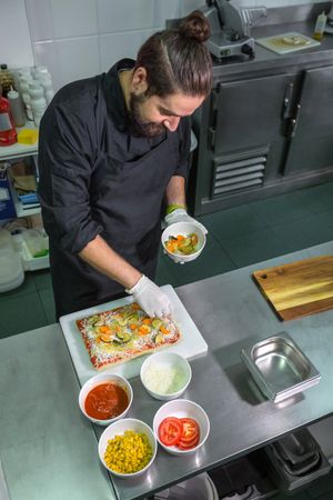 Professional male chef adding fresh healthy ingredients to vegetarian pizza in restaurant kitchen