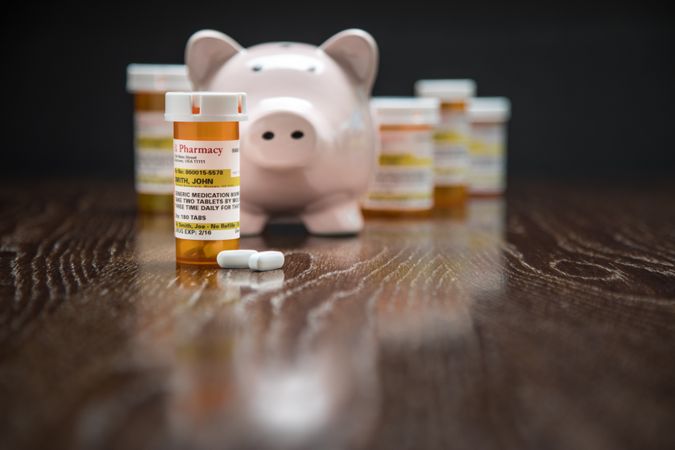Variety of Non-Proprietary Prescription Medicine Bottles, Pills and Piggy Bank on Reflective Wooden Surface.