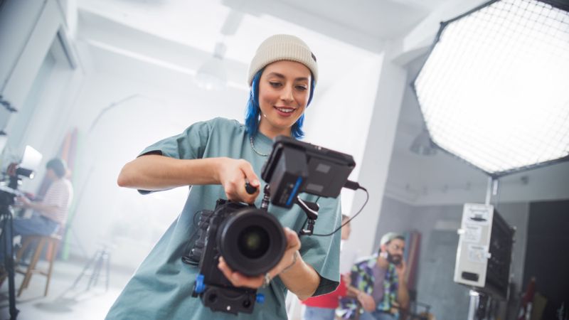 Young Female Filmmaker In A Beanie Adjusts A Camera On A Busy Film Set, Capturing The Essence Of Modern Cinematography With Her Team In The Background, Focused On A Creative Project.