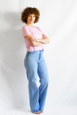 Full body of woman with curly hair standing against studio wall crossing arms