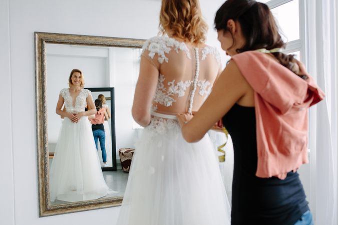 Reflection in mirror of female trying on wedding dress in a shop with women assistant