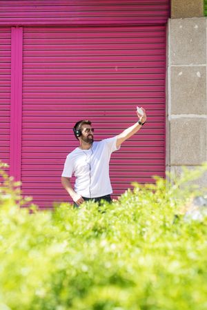 Man in headphones taking selfie with phone in front of pink wall