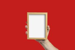 Hand holding an empty wooden picture frame mockup on a red background.