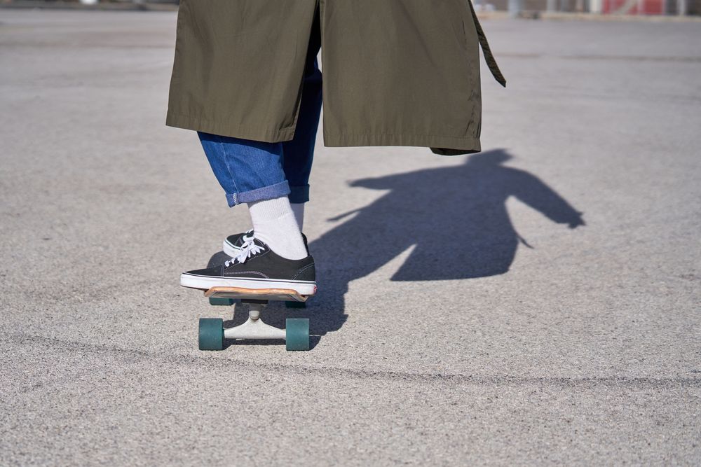 Crop man in sneakers and raincoat riding skateboard