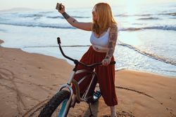 Cheerful woman taking selfie on smartphone while resting with bicycle at sunny shore