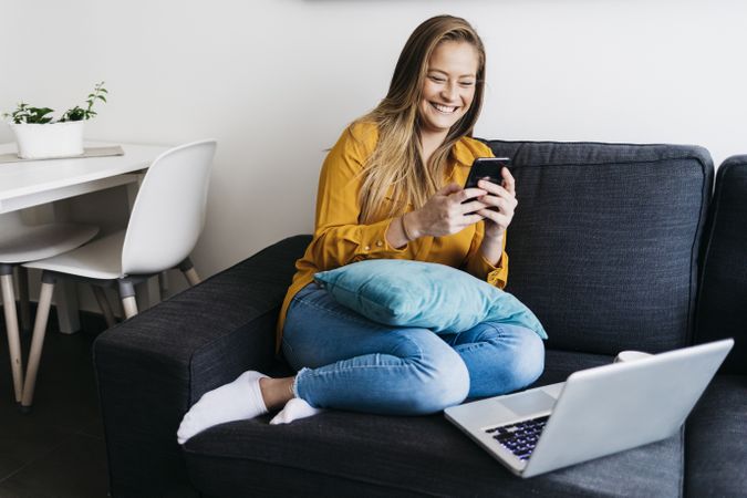 Smiling long haired female sitting on sofa with laptop computer working remotely from home checking phone