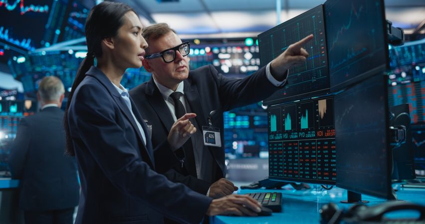 Team of Two Multiethnic Stock Exchange Brokers Using Computer, Chatting About the Asset Management Strategy for an International Business Client, Working in an Office Building with Computer Screens