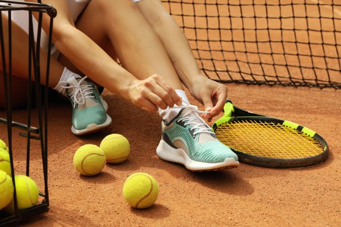 Woman tying shoelaces on clay court with racket and tennis balls