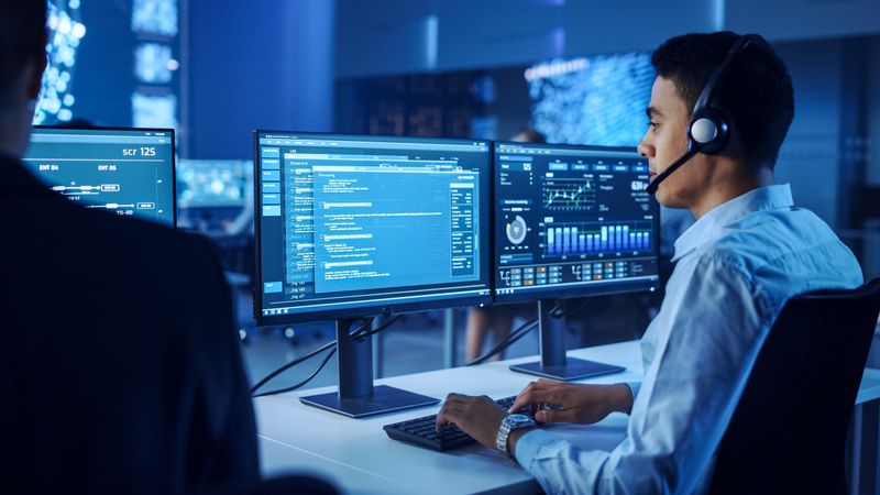 Professional Male Monitors Cybersecurity Data on Dual Screens in a High-tech Control Room, Optimizing Strategies With Precision and Innovation.
