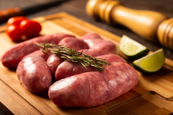 Raw sausages arranged on wooden board with rosemary, lime and tomatoes