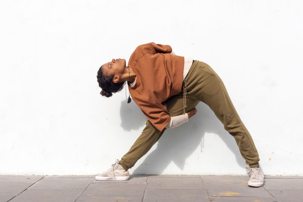 Stylish young female runner in athletic clothes stretching before running against wall